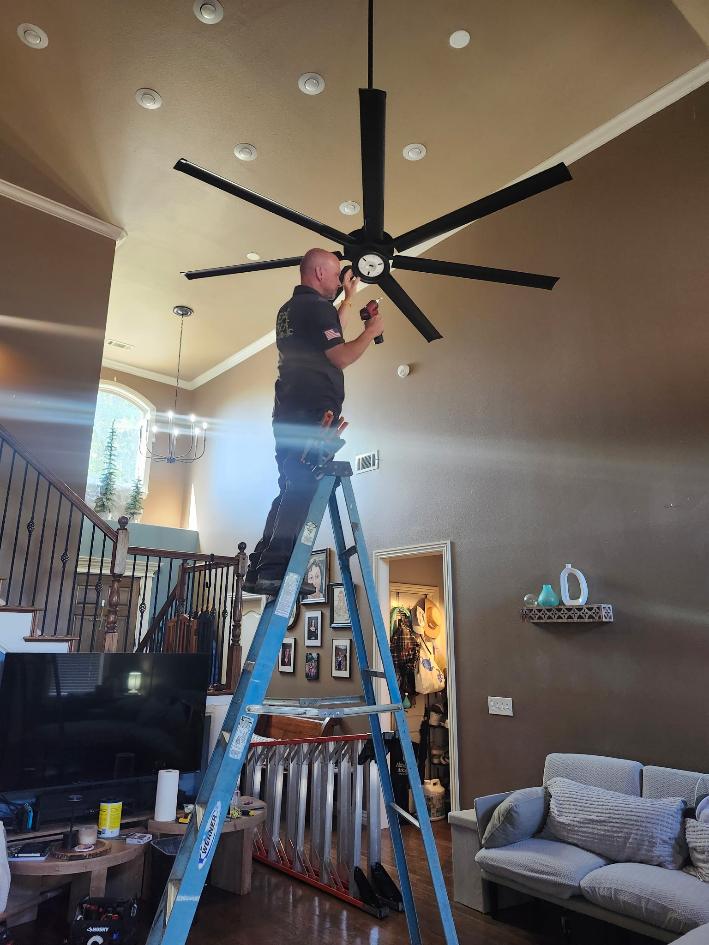 Technician on a ladder working beneath a large black ceiling fan in a living room